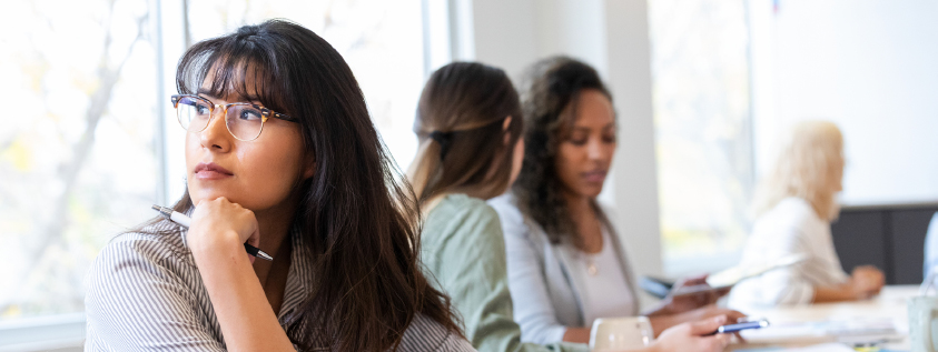 A thoughtful young woman with glasses sits in a classroom, resting her chin on her hand while looking away. In the background, two other women are engaged in work at a table, and another woman is seated nearby, looking at papers.