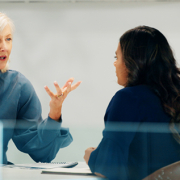 Two women are sitting at a table in an office setting, engaged in a discussion. One woman has short grey hair and is gesturing with her hands, while the other woman, with long dark hair, is listening attentively.