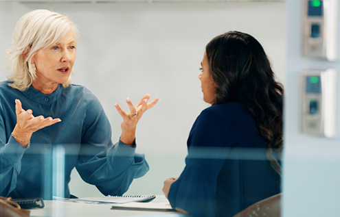 Two women are sitting at a table in an office setting, engaged in a discussion. One woman has short grey hair and is gesturing with her hands, while the other woman, with long dark hair, is listening attentively.