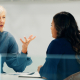 Two women are sitting at a table in an office setting, engaged in a discussion. One woman has short grey hair and is gesturing with her hands, while the other woman, with long dark hair, is listening attentively.