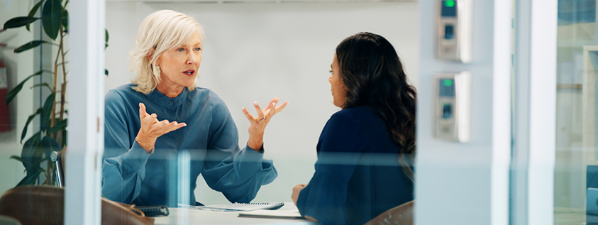 Two women are sitting at a table in an office setting, engaged in a discussion. One woman has short grey hair and is gesturing with her hands, while the other woman, with long dark hair, is listening attentively.