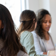 Autistic burnout in adults A thoughtful young woman with glasses sits in a classroom, resting her chin on her hand while looking away. In the background, two other women are engaged in work at a table, and another woman is seated nearby, looking at papers.