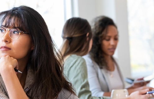 A thoughtful young woman with glasses sits in a classroom, resting her chin on her hand while looking away. In the background, two other women are engaged in work at a table, and another woman is seated nearby, looking at papers.
